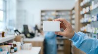 Scientist in blue lab coat holding blank white card in laboratory with shelves of chemicals and equipment in background, suitable for healthcare, research, and medical professional concepts.