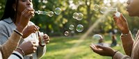 Young diverse women blowing soap bubbles outdoors in sunny park setting, enjoying leisure time together with playful summer activity and friendship bonding moment.