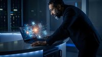 Focused businessman working on laptop with holographic digital interface icons in modern office environment for technology and innovation concepts.