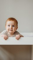 Adorable redhead baby with blue eyes in striped outfit standing at white table against neutral background for family and childcare concepts.