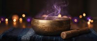 Tibetan singing bowl with wooden mallet on dark blue fabric creating mystical purple smoke against warm bokeh lights for meditation and wellness practices.