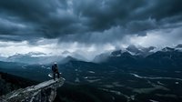 Hiker standing on rocky mountain peak overlooking dramatic valley landscape with stormy clouds and snow-capped peaks, adventure and outdoor exploration.