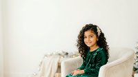 Young woman with long curly dark hair and medium skin tone wearing emerald green velvet jacket, seated in cream armchair against neutral background, smiling at camera for portrait photography.