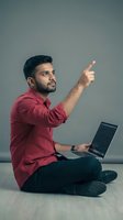 Young Indian businessman in red shirt pointing while using laptop computer sitting on floor for presentation or remote work concept.