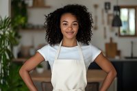 Young African American woman with curly hair wearing white t-shirt and beige apron smiling confidently in modern kitchen interior with natural lighting and plants.