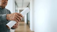 Senior man with age spots and wrinkles holding rolled diploma certificate in hallway. Mature male displaying graduation or achievement document indoors.