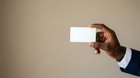 Male professional with dark skin tone wearing navy business suit holding blank white business card against neutral beige background for corporate branding mockup.