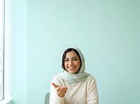 Young woman wearing cream-colored hijab and beige long-sleeve top gestures welcomingly against soft mint green background, suitable for diversity, inclusion, and lifestyle marketing campaigns.
