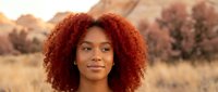 Young African American woman with curly red hair smiling outdoors during golden hour sunset in natural landscape for lifestyle and beauty concepts.