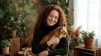 Young woman with curly red hair embracing orange tabby cat in cozy plant-filled home interior. Pet owner bonding with feline companion indoors.