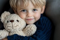 Smiling young Caucasian boy with blonde hair and blue eyes hugging soft beige teddy bear, wearing navy blue knit sweater against dark background.