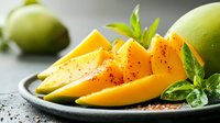 Fresh ripe mango slices with green basil leaves and spices on dark plate, selective focus on juicy tropical fruit with natural lighting.