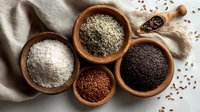 Assortment of organic seeds and grains in wooden bowls including white rice, hemp seeds, black quinoa and red flax seeds on neutral linen background.