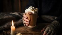 Cozy winter evening with hands holding warm hot chocolate mug topped with whipped cream and cinnamon, soft candlelight creating intimate atmosphere for comfort drinks.