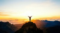 Person with arms raised in victory standing on mountain peak at sunset, celebrating achievement and success with golden sky backdrop.