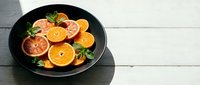 Sliced citrus fruits arranged on black plate with fresh mint garnish, overhead view on white wooden surface for food styling and culinary content.