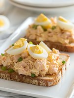 Open-faced tuna salad sandwich with sliced hard-boiled eggs and fresh chives on whole grain bread served on white plate for healthy lunch meal.