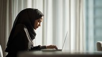 Young Middle Eastern woman in hijab working on laptop by window with soft natural light. Professional female entrepreneur in traditional attire focused on digital task.