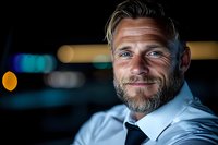 Confident mature Caucasian businessman with gray beard wearing white shirt and tie smiles warmly against dark background with bokeh lights. Professional portrait.