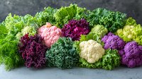 Fresh colorful cauliflower and kale varieties arranged in vibrant display showing pink, purple, white and green cruciferous vegetables with curly lettuce and herbs.
