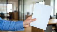 Male professional in blue shirt holding blank white card or document in modern office environment with blurred background for mockup and branding purposes.