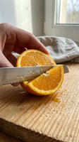 Hand holding fresh orange slice being cut with knife on wooden cutting board in bright kitchen with window light.