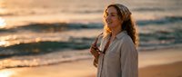 Woman with long curly hair in white linen shirt standing on beach at sunset, holding sunglasses, ocean waves in background.