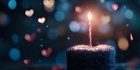 Chocolate birthday cake with single lit candle against dreamy bokeh background with heart-shaped lights creating magical celebration atmosphere.