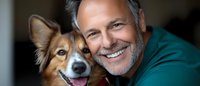 Mature Caucasian man with gray hair and beard wearing green sweater smiling while hugging happy mixed breed dog with pointed ears in close-up portrait.