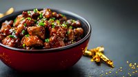 Glazed chicken pieces in red bowl garnished with sesame seeds and green onions, served on dark background with golden spices scattered around.