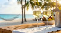 Two glasses of chilled white wine on wooden table at tropical beach resort during golden hour sunset with palm trees and turquoise ocean in background.