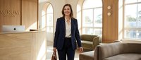 Professional businesswoman in navy suit walking through modern office lobby with natural lighting and contemporary furniture for corporate presentations.