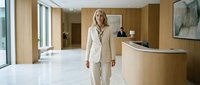 Professional blonde businesswoman in elegant beige suit walking through modern corporate office lobby with curved reception desk and contemporary interior design.