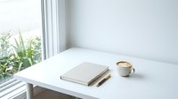 Minimalist workspace with closed notebook, pen, and coffee cup on white desk near bright window with plant view.