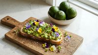 Avocado toast topped with edible flowers and microgreens on wooden cutting board, fresh avocados in bowl, healthy brunch food styling for culinary and wellness content.