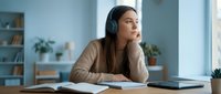 Young woman with dark hair and South Asian ethnicity wearing headphones, sitting at desk with notebook and tablet, appearing focused and contemplative while studying or working remotely in bright modern office space.