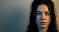 Young brunette woman with intense blue eyes and natural makeup looking directly at camera with serious expression against soft blurred background for portrait photography.