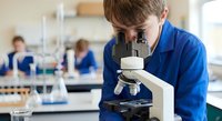 Young male student in blue lab coat using microscope in science laboratory classroom with classmates working in background.