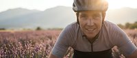 Smiling mature Caucasian male cyclist in helmet riding through lavender field at golden hour sunset with mountains in background for outdoor recreation.