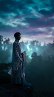 Young Asian man in traditional robe standing on cliff edge gazing at dramatic twilight sky with purple and teal clouds for meditation and spiritual themes.