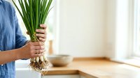 Young adult male with medium skin tone holding fresh green onions with roots and soil visible, wearing casual blue shirt in bright kitchen setting with wooden countertop and white background for culinary and gardening content.