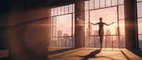 Silhouette of businessman standing at large office windows during golden hour sunset with city skyline view, arms outstretched in success gesture.