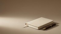 Closed cream hardcover notebook with bookmark ribbon resting on dark wooden surface against neutral beige background, suitable for stationery, education, and publishing design projects.