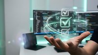Businessman hand holding digital checkmarks and data visualization with computer screens in background, representing quality assurance, task completion, and software development workflow.