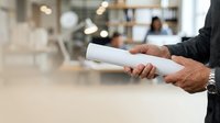 Mature male professional holding rolled blueprint in modern office environment with colleagues working in background.