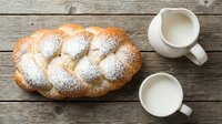 Golden braided challah bread dusted with powdered sugar alongside white ceramic mugs filled with fresh milk on rustic wooden table for breakfast bakery.