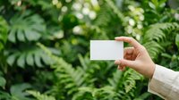 Hand holding blank white card against lush green fern foliage background. Ideal for business cards, eco-friendly branding, nature-based marketing materials, and environmental campaigns.