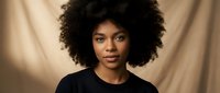 Young African American woman with natural curly afro hair wearing black top against neutral beige background for portrait photography and lifestyle content.