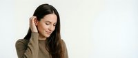 Young woman with long dark hair and olive complexion wearing olive-toned sweater, touching her ear with gentle expression against white background for portrait and lifestyle use.