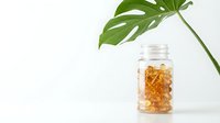 Transparent glass jar filled with golden amber gel capsules next to fresh green monstera leaf on white background, isolated for wellness and supplement product design use.